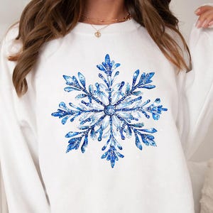 Könnte beinhalten: Weißes Sweatshirt mit einem großen, detaillierten blauen Schneeflocken-Design. Die Schneeflocke hat komplizierte Muster und einen Aquarelleffekt mit Akzenten in Hellblau und Weiß. Das Sweatshirt ist langärmlig und hat einen Rundhalsausschnitt.