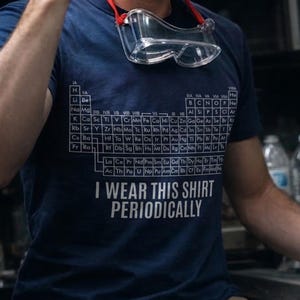 May include: Navy blue t-shirt featuring the periodic table of elements and the text "I WEAR THIS SHIRT PERIODICALLY" in white. Clear safety glasses with a red strap are hanging on the shirt.