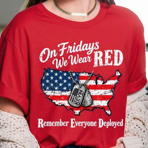 May include: A red t-shirt featuring the white text On Fridays We Wear RED and Remember Everyone Deployed. The graphic includes a distressed United States map filled with the American flag and two silver dog tags, one labeled KEVIN.