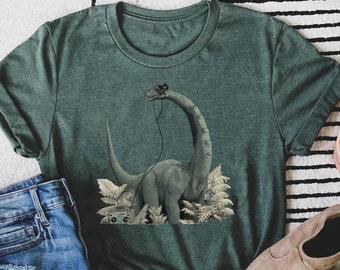 Può includere: Una maglietta verde con una grafica di un dinosauro che indossa delle cuffie e tiene un boombox. Il dinosauro è in piedi in un ambiente di giungla.