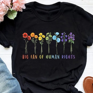 Könnte beinhalten: Schwarzes T-Shirt mit einem bunten Blumendesign und dem Text "BIG FAN OF HUMAN RIGHTS" in Regenbogenfarben. Die Blumen umfassen rote Mohnblumen, orangefarbene Ringelblumen, gelbe Gänseblümchen und blaue Glockenblumen.