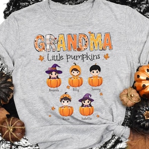 May include: Grey t-shirt with the text "Grandma Little Pumpkins" in orange and brown. Five cartoon children dressed as pumpkins and witches are arranged in a row below the text. Each child is labelled with a name: Amanda, Billy, Tommy, Kevin, and Isabella.