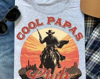 May include: A grey crew neck t-shirt with a cowboy on horseback, holding a gun, against a sunset. The text "COOL PAPAS Club" is printed in red, vintage style. Sunglasses and a brown cap are also visible in the image.