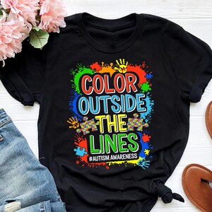 Puede incluir: Camiseta negra con un diseño gráfico colorido que dice "Color Outside The Lines" y "#Autism Awareness".