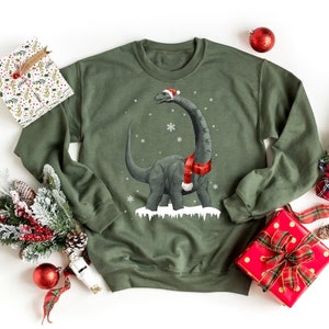 Peut inclure: Un sweat-shirt de Noël vert avec un dinosaure de dessin animé portant un bonnet de Père Noël et une écharpe rouge. Le dinosaure est debout dans la neige avec des flocons de neige qui tombent autour de lui.