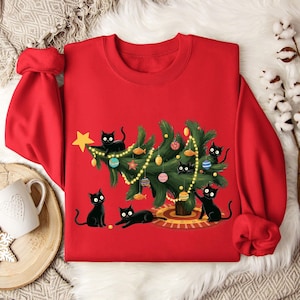 Op de afbeelding: Rode sweatshirt met een kerstboomontwerp versierd met ornamenten en lichtjes, omringd door illustraties van zwarte katten. Een gouden ster staat bovenop de boom. De katten staan in verschillende poses, wat een speels vakantiethema cre&euml;ert.
