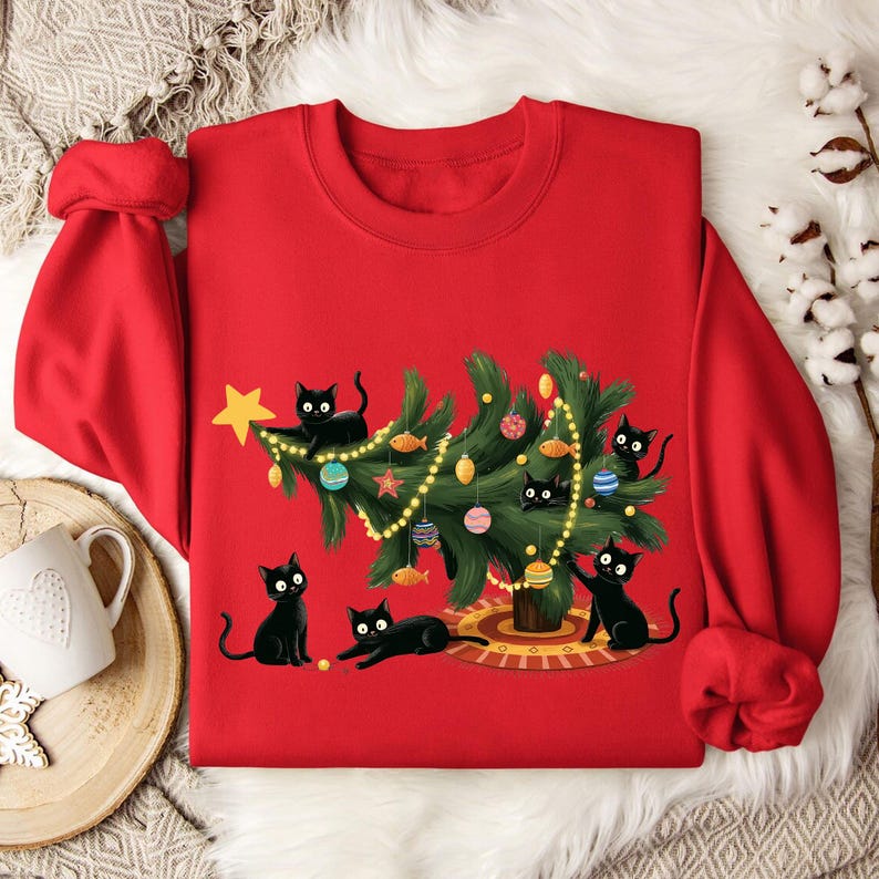 Op de afbeelding: Rode sweatshirt met een kerstboomontwerp versierd met ornamenten en lichtjes, omringd door illustraties van zwarte katten. Een gouden ster staat bovenop de boom. De katten staan in verschillende poses, wat een speels vakantiethema cre&euml;ert.
