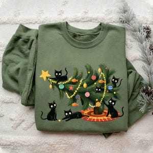 Op de afbeelding: Olijfgroene sweatshirt met een kerstboomontwerp. De boom is versierd met ballen en lichtjes, omringd door zwarte katten. De sweatshirt heeft een ronde hals en lange mouwen, perfect voor de wintermaanden.