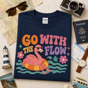 May include: A navy blue crew neck t-shirt featuring a graphic of a pink flamingo wearing heart-shaped sunglasses while floating on an orange and yellow inner tube, with the text "GO WITH THE FLOW" in orange, pink, and purple wavy lettering.