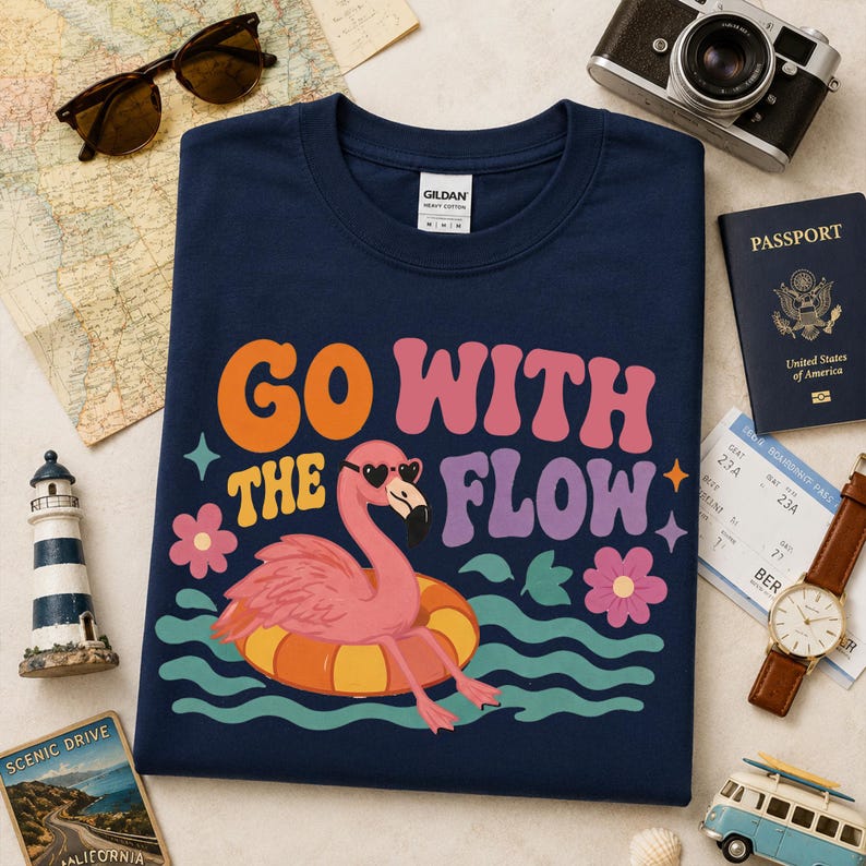 May include: A navy blue crew neck t-shirt featuring a graphic of a pink flamingo wearing heart-shaped sunglasses while floating on an orange and yellow inner tube, with the text "GO WITH THE FLOW" in orange, pink, and purple wavy lettering.