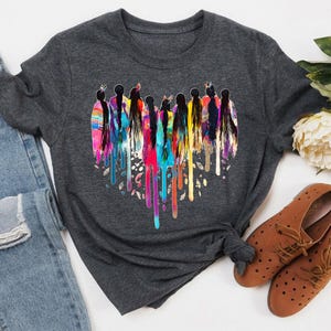 May include: A dark gray t-shirt with a colorful graphic print of a group of women standing in a heart shape. The women are wearing traditional Native American clothing and have long, flowing hair. The graphic is dripping with colorful paint.