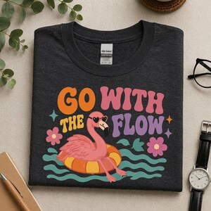 May include: A dark charcoal gray crew neck t-shirt featuring a graphic of a pink flamingo wearing heart-shaped sunglasses while floating in an orange and yellow inner tube. The text reads GO WITH THE FLOW in orange, pink, and purple retro lettering.