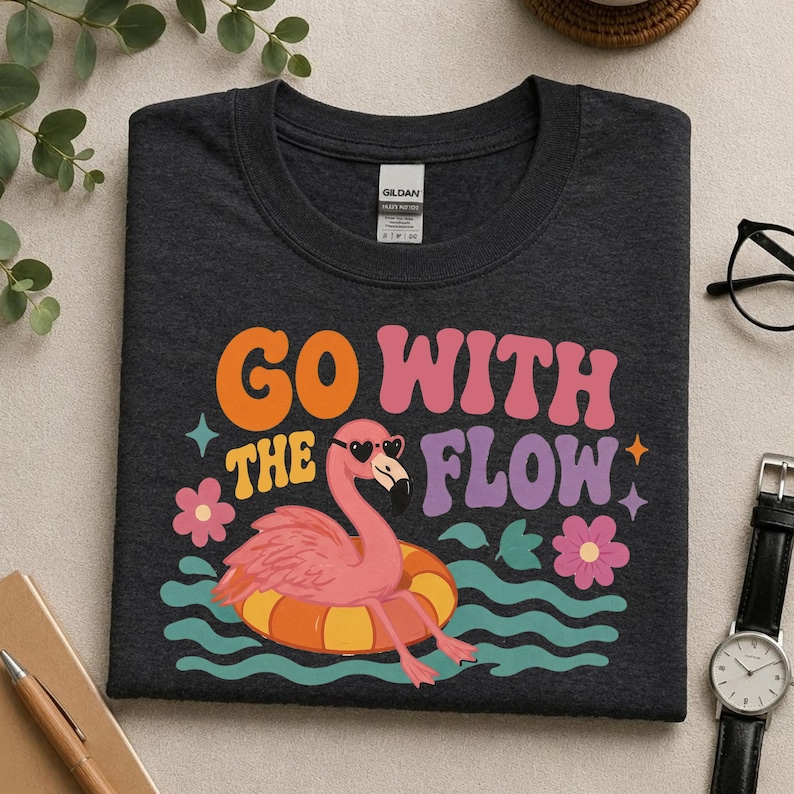 May include: A dark charcoal gray crew neck t-shirt featuring a graphic of a pink flamingo wearing heart-shaped sunglasses while floating in an orange and yellow inner tube. The text reads GO WITH THE FLOW in orange, pink, and purple retro lettering.