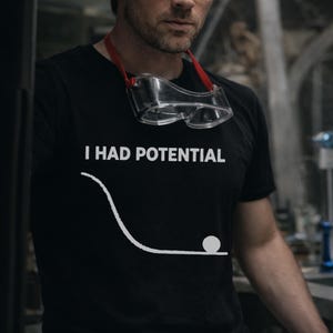 Puede incluir: Camiseta negra con el texto "I HAD POTENTIAL" y un gráfico blanco de una bola en una trayectoria curva. Gafas de seguridad transparentes con una correa roja alrededor del cuello.