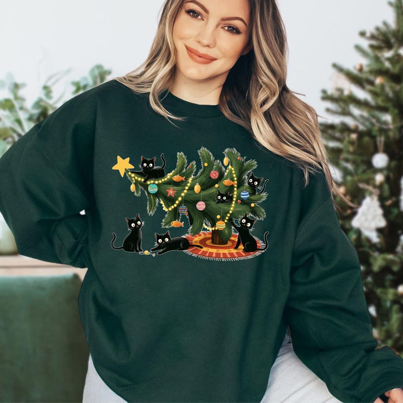 Op de afbeelding: Donkergroene sweatshirt met een kerstboomontwerp. Het heeft een kerstboom versierd met ballen en lichtjes, omringd door illustraties van zwarte katten. De katten staan in verschillende poses. Een gouden ster staat bovenop de boom.