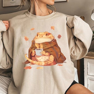 May include: A beige sweatshirt with a graphic of a bearded dragon wearing a blanket and holding a cup of coffee.  Autumn leaves are scattered around the dragon.