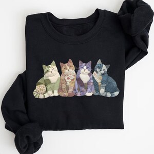Peut inclure: Sweat-shirt noir orn&eacute; d'une rang&eacute;e de cinq chats souriants en appliqu&eacute;. Chaque chat pr&eacute;sente un motif patchwork unique dans les tons de vert, marron, violet et bleu. Les chats sont assis, les yeux ferm&eacute;s, cr&eacute;ant un design confortable et fantaisiste.