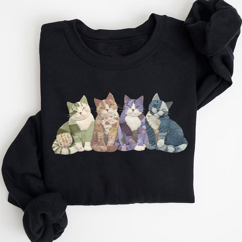Peut inclure: Sweat-shirt noir orn&eacute; d'une rang&eacute;e de cinq chats souriants en appliqu&eacute;. Chaque chat pr&eacute;sente un motif patchwork unique dans les tons de vert, marron, violet et bleu. Les chats sont assis, les yeux ferm&eacute;s, cr&eacute;ant un design confortable et fantaisiste.