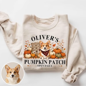 Könnte beinhalten: Ein cremefarbenes Sweatshirt mit Rundhalsausschnitt und einem Corgi-Motiv, der ein rot-schwarz kariertes Halstuch trägt. Das Design zeigt Kürbisse, Herbstblätter, ein Holzschild und den Text OLIVER'S PUMPKIN PATCH OPEN DAILY.