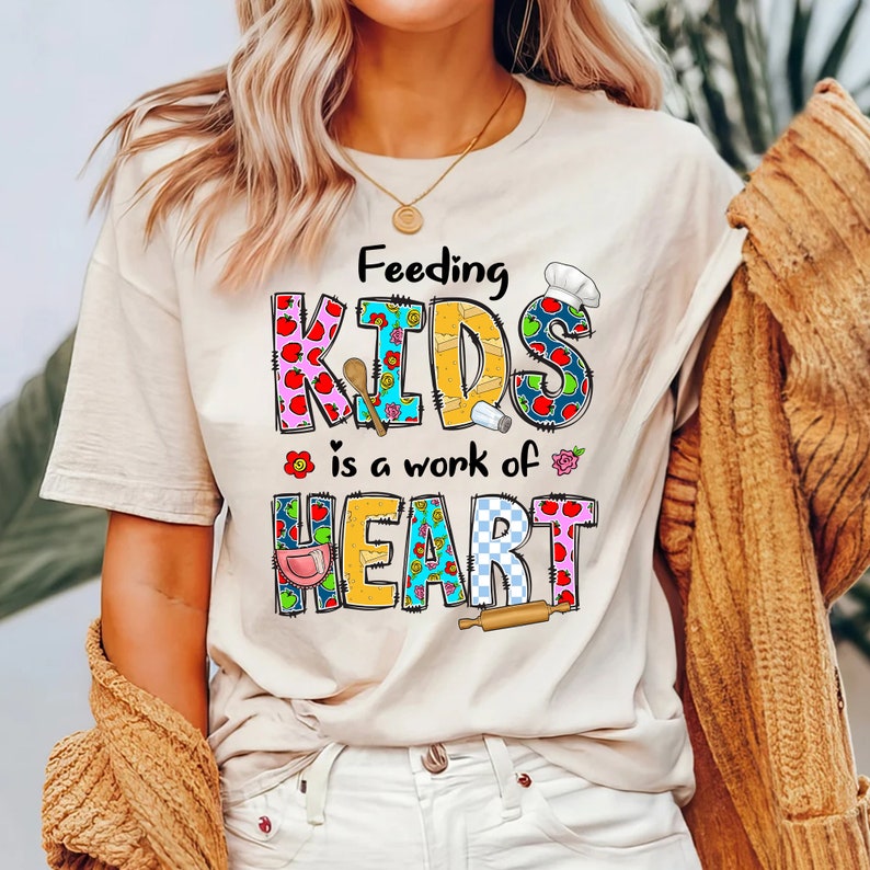 May include: A beige t-shirt with a colorful graphic that says "Feeding Kids is a work of heart". The graphic features a chef's hat, apples, and other kitchen-related imagery.
