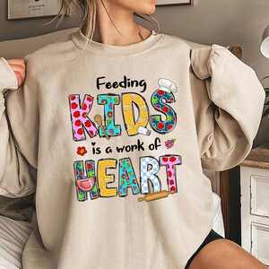 May include: Beige sweatshirt with a colorful graphic that says "Feeding Kids is a work of heart". The graphic is made up of patchwork letters and includes images of a chef's hat, apples, a rolling pin, a salt shaker, and a wooden spoon.