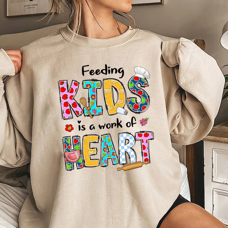 May include: Beige sweatshirt with a colorful graphic that says "Feeding Kids is a work of heart". The graphic is made up of patchwork letters and includes images of a chef's hat, apples, a rolling pin, a salt shaker, and a wooden spoon.