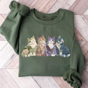 Peut inclure: Un sweat-shirt vert olive avec une rang&eacute;e de quatre illustrations de chats souriants. Chaque chat a un design patchwork unique dans les tons de vert, orange, violet et bleu. Le sweat-shirt a des manches longues avec des poignets c&ocirc;tel&eacute;s.