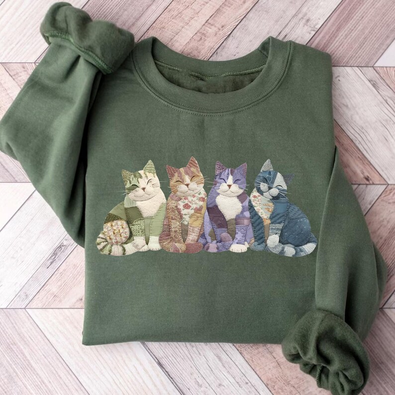 Peut inclure: Un sweat-shirt vert olive avec une rang&eacute;e de quatre illustrations de chats souriants. Chaque chat a un design patchwork unique dans les tons de vert, orange, violet et bleu. Le sweat-shirt a des manches longues avec des poignets c&ocirc;tel&eacute;s.