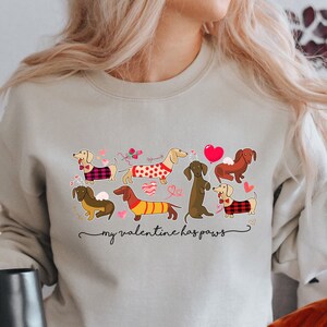Puede incluir: Una sudadera beige con un gr&aacute;fico de seis perros salchicha con diversos su&eacute;teres y posturas, rodeados de peque&ntilde;os corazones y flores rojas y rosas. El texto debajo de los perros dice "my valentine has paws" en letra cursiva negra.