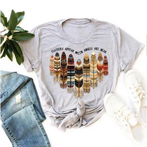 May include: A gray t-shirt with a graphic of colorful feathers and the text "Feathers appear when angels are near".