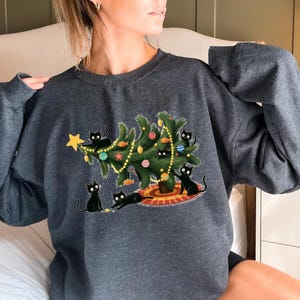 Op de afbeelding: Donkergrijze sweatshirt met een kerstboomontwerp, versierd met kleurrijke ballen en lichtslingers. Zwarte katten zijn rond de boom afgebeeld, sommigen spelen. Een gouden ster staat bovenop de boom. Een feestelijk en gezellig ontwerp.