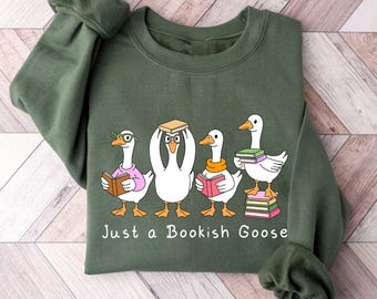 Puede incluir: Sudadera verde con un gráfico de cuatro gansos de dibujos animados, cada uno sosteniendo libros. Los gansos llevan gafas y bufandas. El texto "Just a Bookish Goose" está impreso debajo. La sudadera es de material suave.