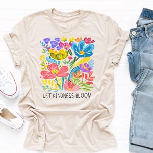 May include: A beige t-shirt with a colorful floral design and the text "Let Kindness Bloom".