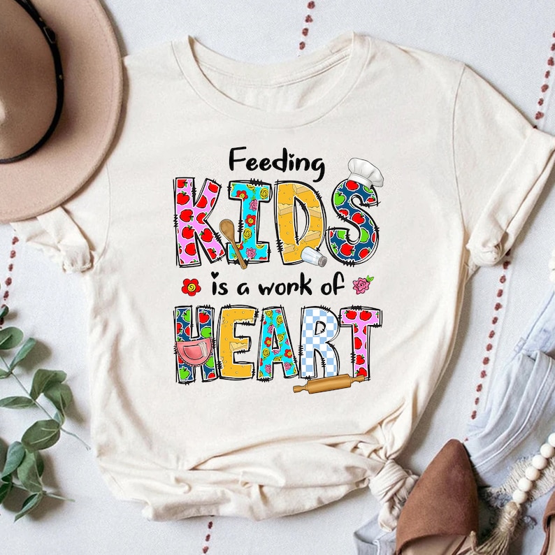May include: A white t-shirt with the text "Feeding Kids is a work of Heart" in colorful patchwork letters. The letters are decorated with images of food and kitchen utensils.