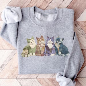 Peut inclure: Un sweat-shirt gris avec un motif de quatre chats souriants de diff&eacute;rentes couleurs et motifs. Les chats sont assis c&ocirc;te &agrave; c&ocirc;te, avec des d&eacute;tails en vert, orange, violet et bleu. Le sweat-shirt a un col rond et des manches longues.