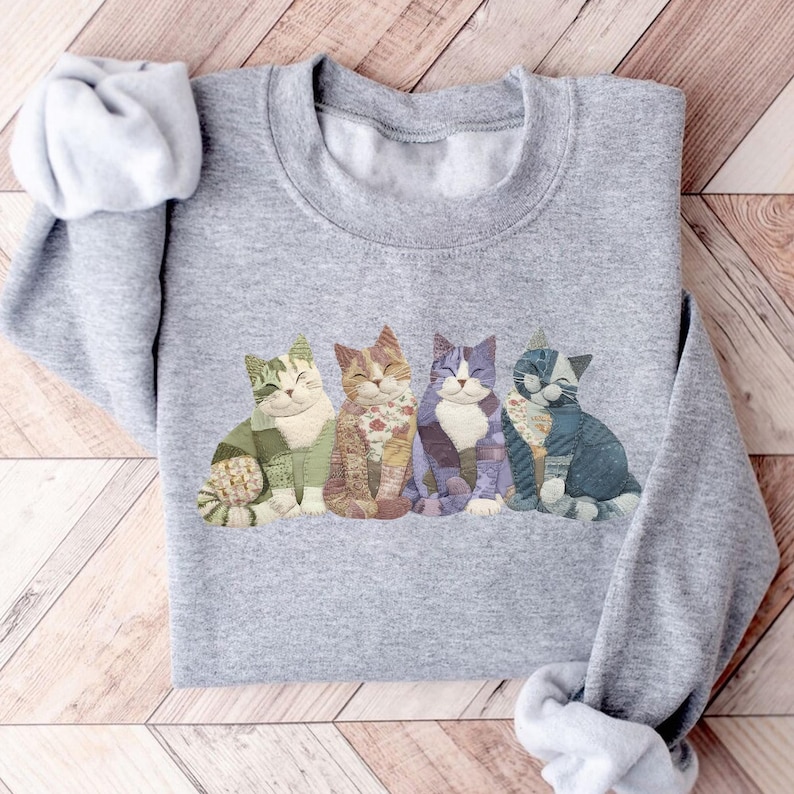 Peut inclure: Un sweat-shirt gris avec un motif de quatre chats souriants de diff&eacute;rentes couleurs et motifs. Les chats sont assis c&ocirc;te &agrave; c&ocirc;te, avec des d&eacute;tails en vert, orange, violet et bleu. Le sweat-shirt a un col rond et des manches longues.