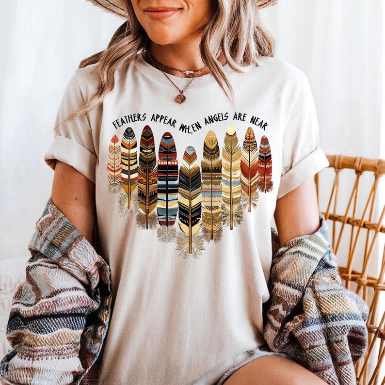 May include: A white t-shirt with a graphic of colorful feathers and the text "Feathers Appear When Angels Are Near".