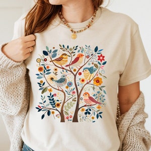 May include: Beige t-shirt featuring a colorful illustration of a tree with birds. The design includes various birds perched on branches with leaves and flowers in shades of blue, yellow, orange, and pink. The shirt is made of a soft material.