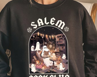 Puede incluir: Sudadera negra con un gráfico blanco de un fantasma leyendo un libro en una habitación espeluznante con el texto "Salem Book Club" en una fuente gótica.