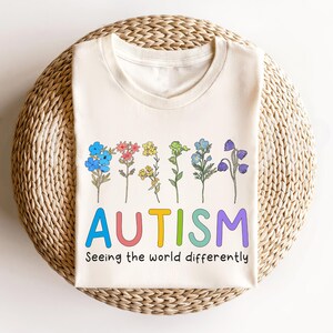May include: A cream-colored t-shirt with a rainbow-colored word "AUTISM" and the text "Seeing the world differently". The word "AUTISM" is surrounded by a row of colorful flowers.