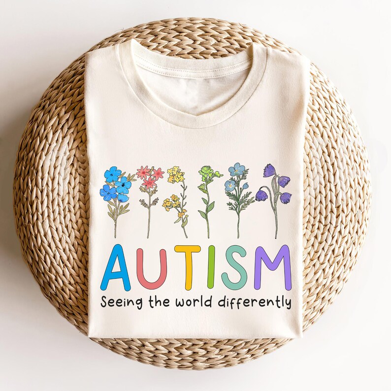 May include: A cream-colored t-shirt with a rainbow-colored word "AUTISM" and the text "Seeing the world differently". The word "AUTISM" is surrounded by a row of colorful flowers.