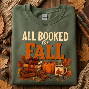 May include: An olive green crewneck sweatshirt featuring the text "ALL BOOKED for FALL" in cream and orange lettering. The graphic includes a stack of three books tied with a red plaid ribbon, a pumpkin, a coffee mug, and eyeglasses.