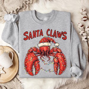 Könnte beinhalten: Ein graues Sweatshirt mit dem Aufdruck "Santa Claws". Darunter ist ein Cartoon-Hummer zu sehen, der einen Weihnachtsmann-Hut und Weihnachtslichter trägt.
