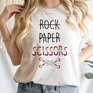 May include: White t-shirt with the text "Rock Paper Scissors" crossed out and replaced with "Scissors" in pink, orange, and white stripes. A pair of scissors with the same color scheme are crossed below the text.