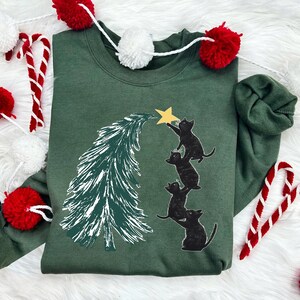 May include: A green Christmas sweatshirt with a black cat design. Four black cats are climbing a green Christmas tree to reach a gold star at the top.