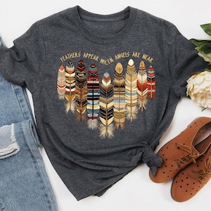 May include: A dark gray t-shirt with a graphic of colorful feathers and the text "Feathers appear when angels are near".