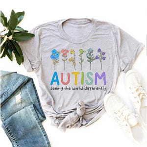 May include: A gray t-shirt with a rainbow-colored flower design and the text "AUTISM Seeing the world differently".