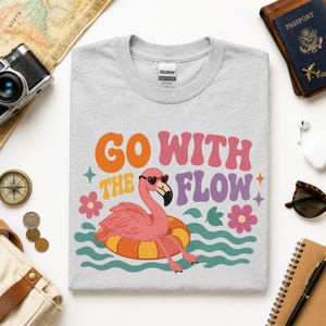 May include: A light gray crew neck t-shirt featuring a graphic of a pink flamingo wearing heart-shaped sunglasses while floating in an orange and yellow inner tube, with the text "GO WITH THE FLOW" in orange, pink, and purple wavy lettering.