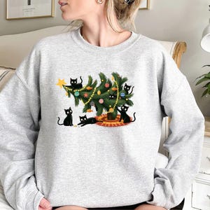 Op de afbeelding: Een lichtgrijs sweatshirt met een kerstboom versierd met ballen en een ster. Zwarte katten spelen rond de boom. Een comfortabele, casual stijl.
