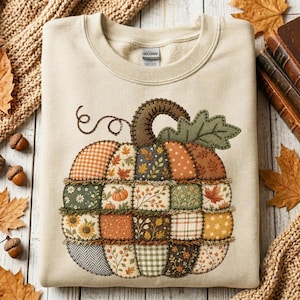 May include: A beige crewneck sweatshirt featuring a large, central pumpkin graphic composed of various fabric-like patches in shades of orange, green, brown, and cream. The pumpkin design includes patterns of gingham, sunflowers, leaves, and berries.